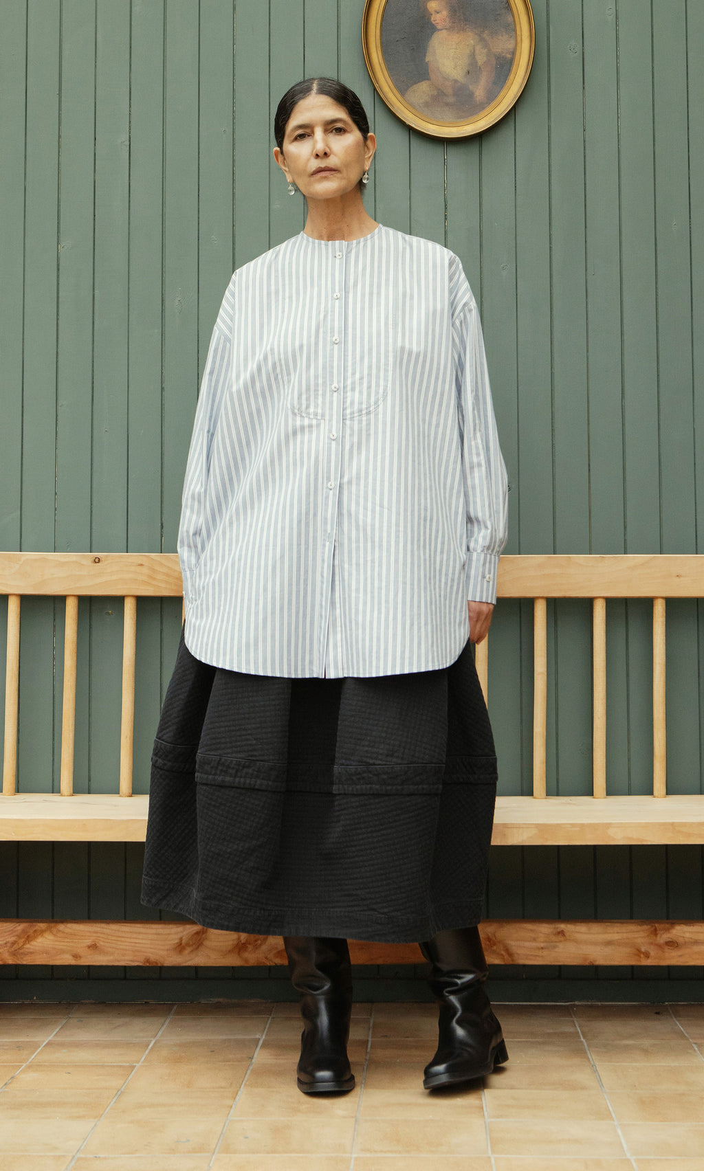 Plümo  Woman in a Tuxedo Shirt and black skirt stands on a wooden bench by a green wall, beneath a framed portrait.