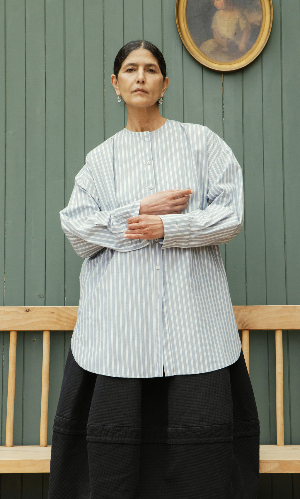 Plümo  Woman in a Tuxedo Shirt and black skirt stands by a wooden bench and green paneled wall with a portrait.