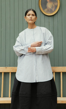 Plümo  Woman in a Tuxedo Shirt and black skirt stands by a wooden bench and green paneled wall with a portrait.
