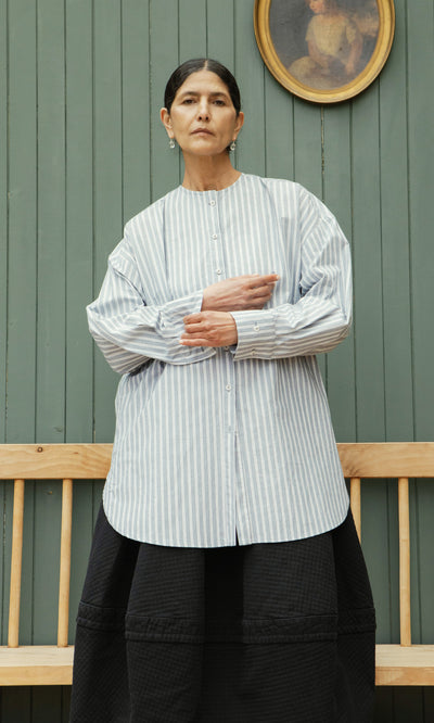 Plümo  Woman in a Tuxedo Shirt and black skirt stands by a wooden bench and green paneled wall with a portrait.