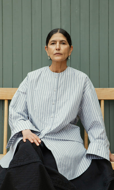 Plümo  Woman in a Tuxedo Shirt sits calmly on a wooden bench against a chambray blue paneled wall.