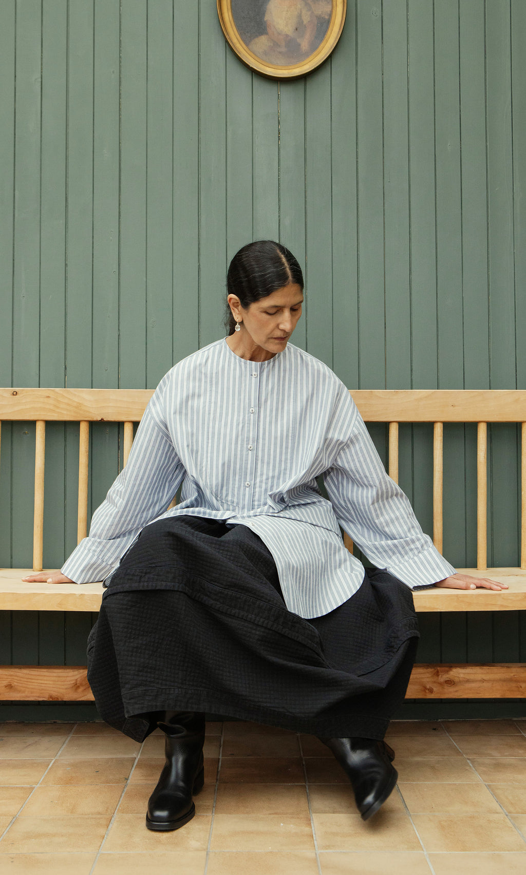 Plümo  Woman in a collarless Tuxedo Shirt and black skirt sits on a wooden bench, looking down, against green paneled wall.