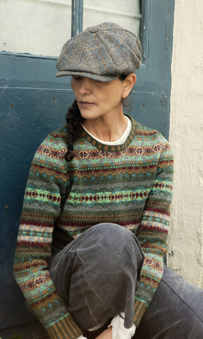 Plümo  Woman in a patterned sweater and Tweed Flat Cap sits by a blue door, her braid draped over one shoulder.