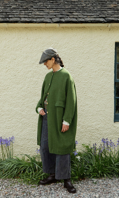 Plümo  Person in a green coat, dark pants, and Tweed Flat Cap stands on gravel near purple flowers and greenery.