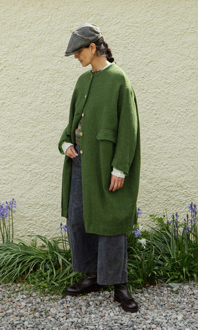 Plümo  Person in a Barra Harris Coat, gray pants, and cap stands on gravel by purple flowers, facing left.