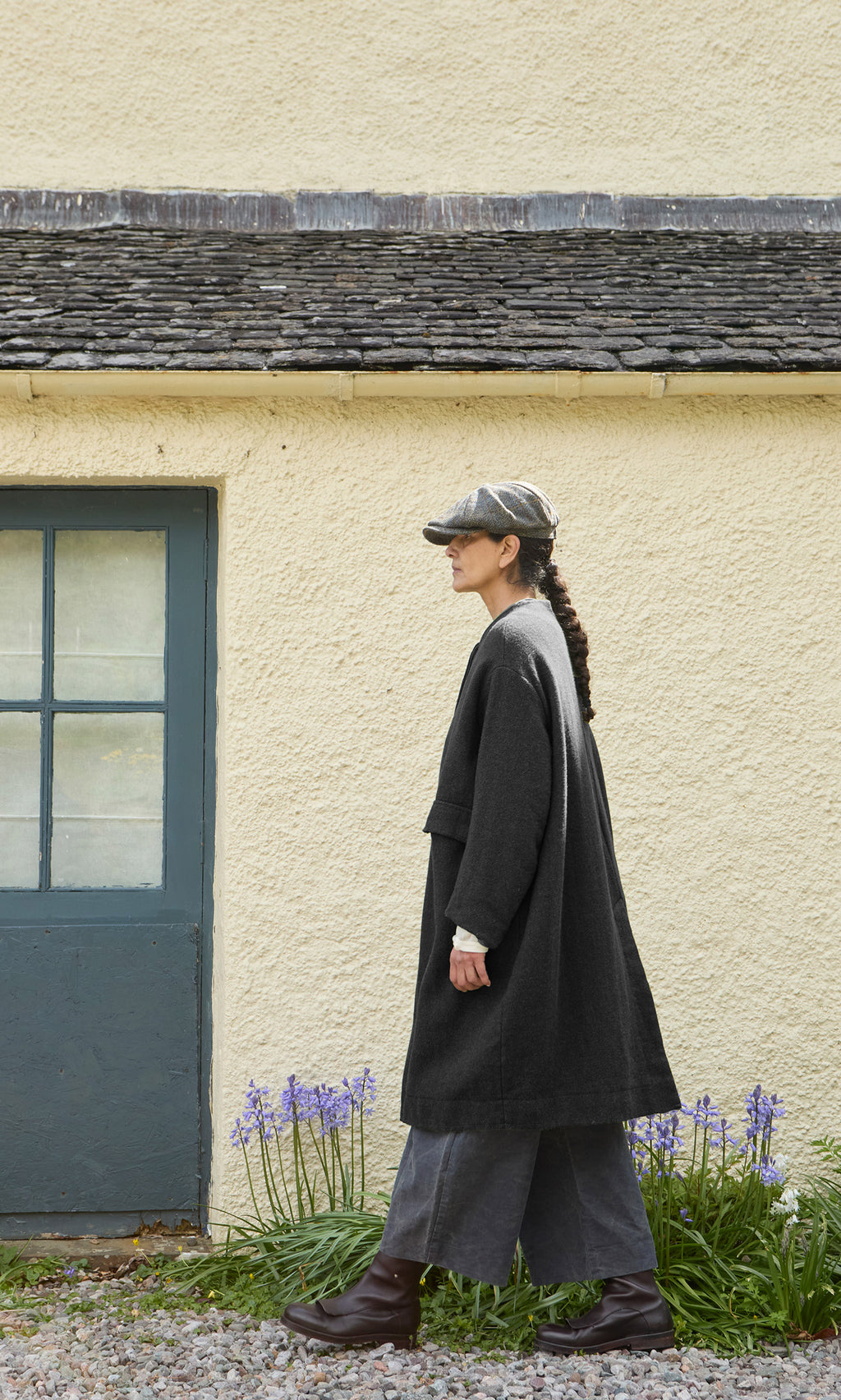 Plümo  A person in a charcoal grey Barra Harris Coat, trousers, and a flat cap walks past a cream-colored building.