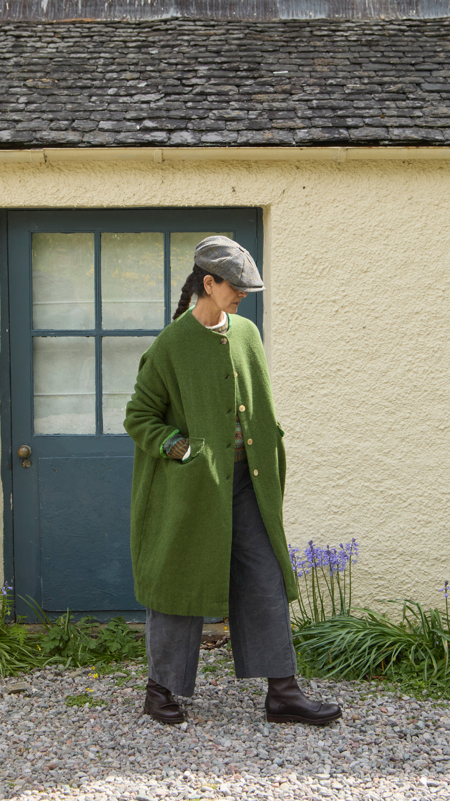 Plümo  A person wearing a Barra Harris Coat, grey wide-leg trousers, and a flat cap stands outside a building.