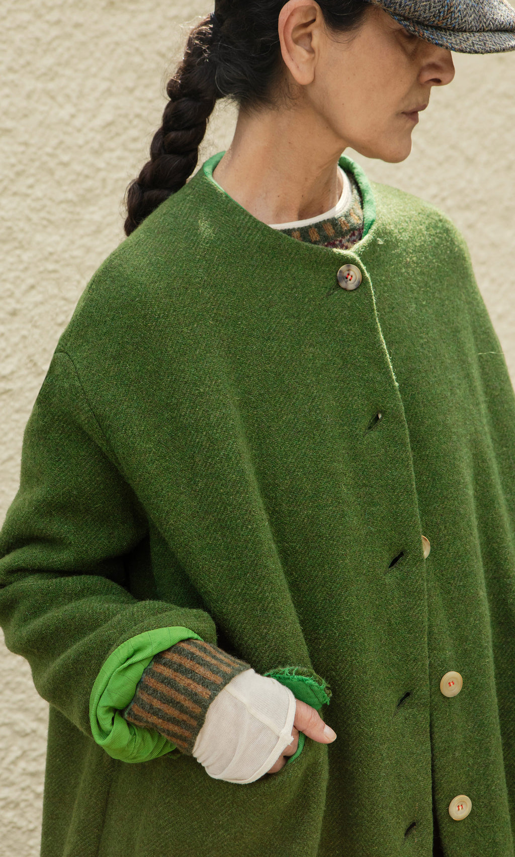 Plümo  Woman in the Barra Harris Coat, with braided hair and cap, stands against a beige wall.