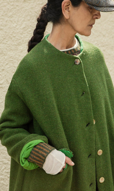 Plümo  Woman in the Barra Harris Coat, with braided hair and cap, stands against a beige wall.