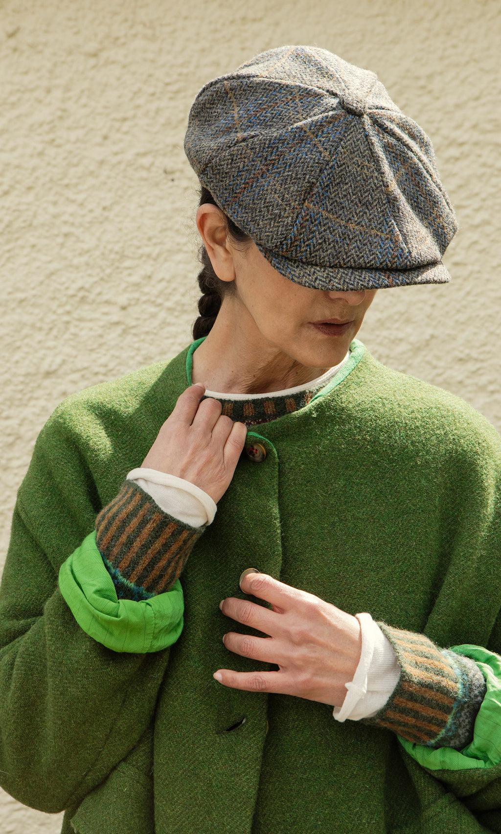 Plümo  Person in a green coat and Tweed Flat Cap looks down, face partly hidden, standing against a textured wall.