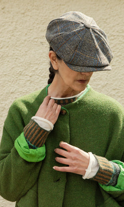 Plümo  Person in a green coat and Tweed Flat Cap looks down, face partly hidden, standing against a textured wall.