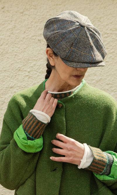 Plümo  Person in a green coat and Tweed Flat Cap looks down, face partly hidden, standing against a textured wall.