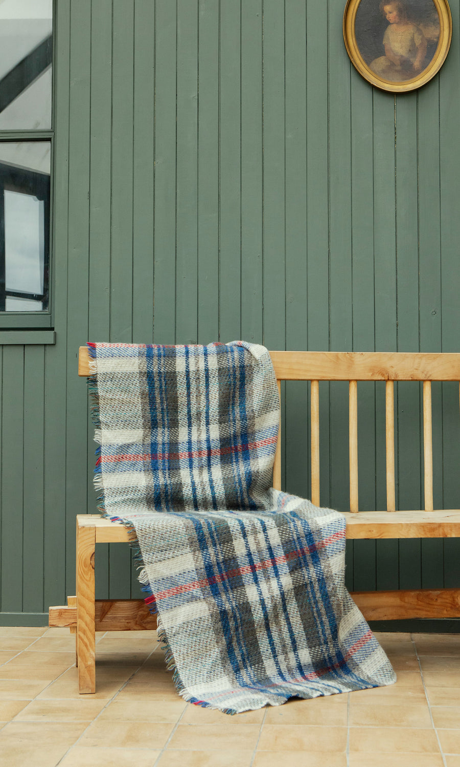 Plümo  Random Throw draped on a wooden bench with a green paneled wall and round portrait above.