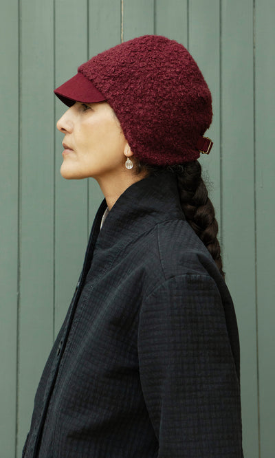 Plümo  A person in a Velo Wool Cap stands in profile before a green wood wall, wearing a dark jacket.