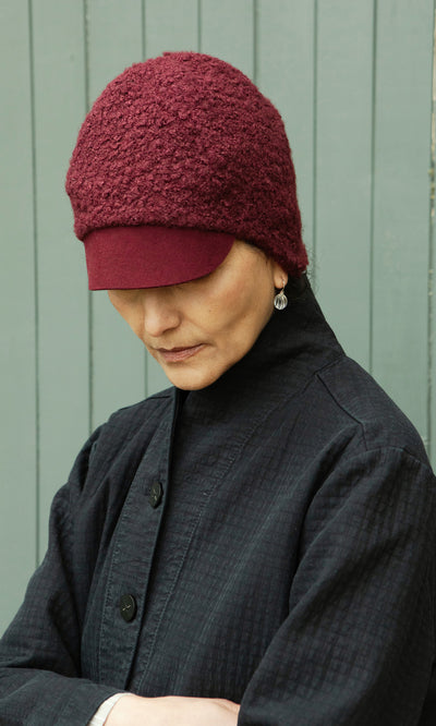 Plümo  Individual in a Velo Wool Cap and black coat looks down, standing before a gray wooden background.