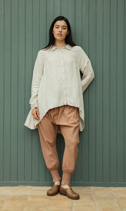 Plümo  Woman in loose cream shirt, Vichy Pantaloons, brown shoes by a green wooden wall.