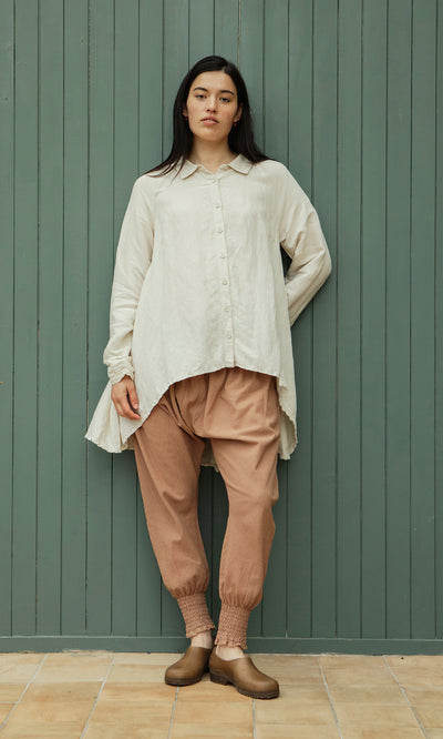 Plümo  Woman in loose cream shirt, Vichy Pantaloons, brown shoes by a green wooden wall.