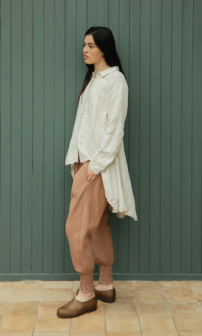 Plümo  Woman in Vichy Pantaloons, loose cream fairtrade cotton top, brown clogs by a green paneled wall.