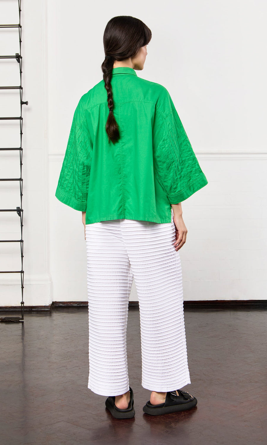 Plümo- Woman with long dark hair in a braid wears Yacco Maricard White Pintucked Pants and a green top indoors.