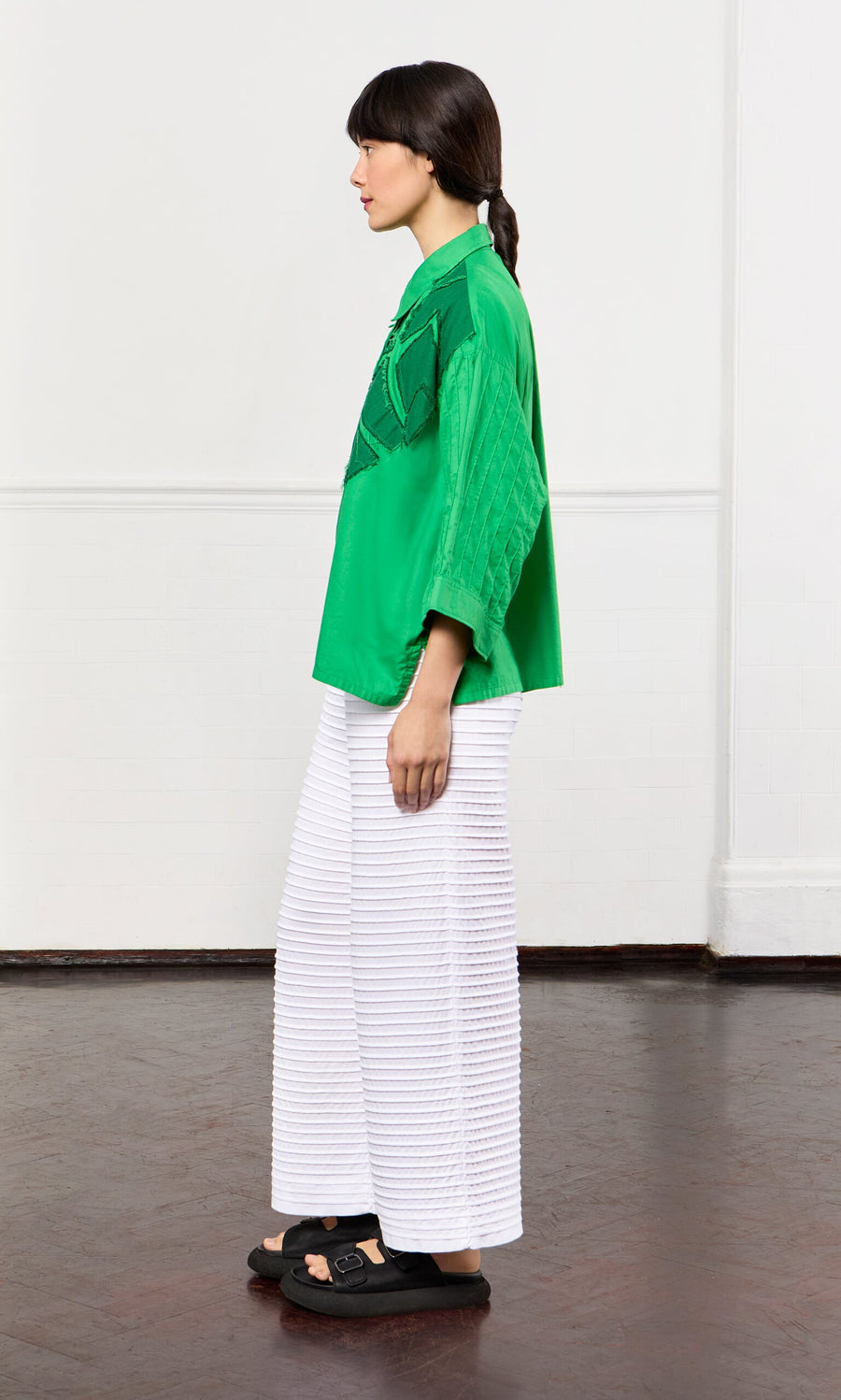 Plümo- A woman in a green shirt and White Pintucked Pants stands sideways on a dark wood floor.