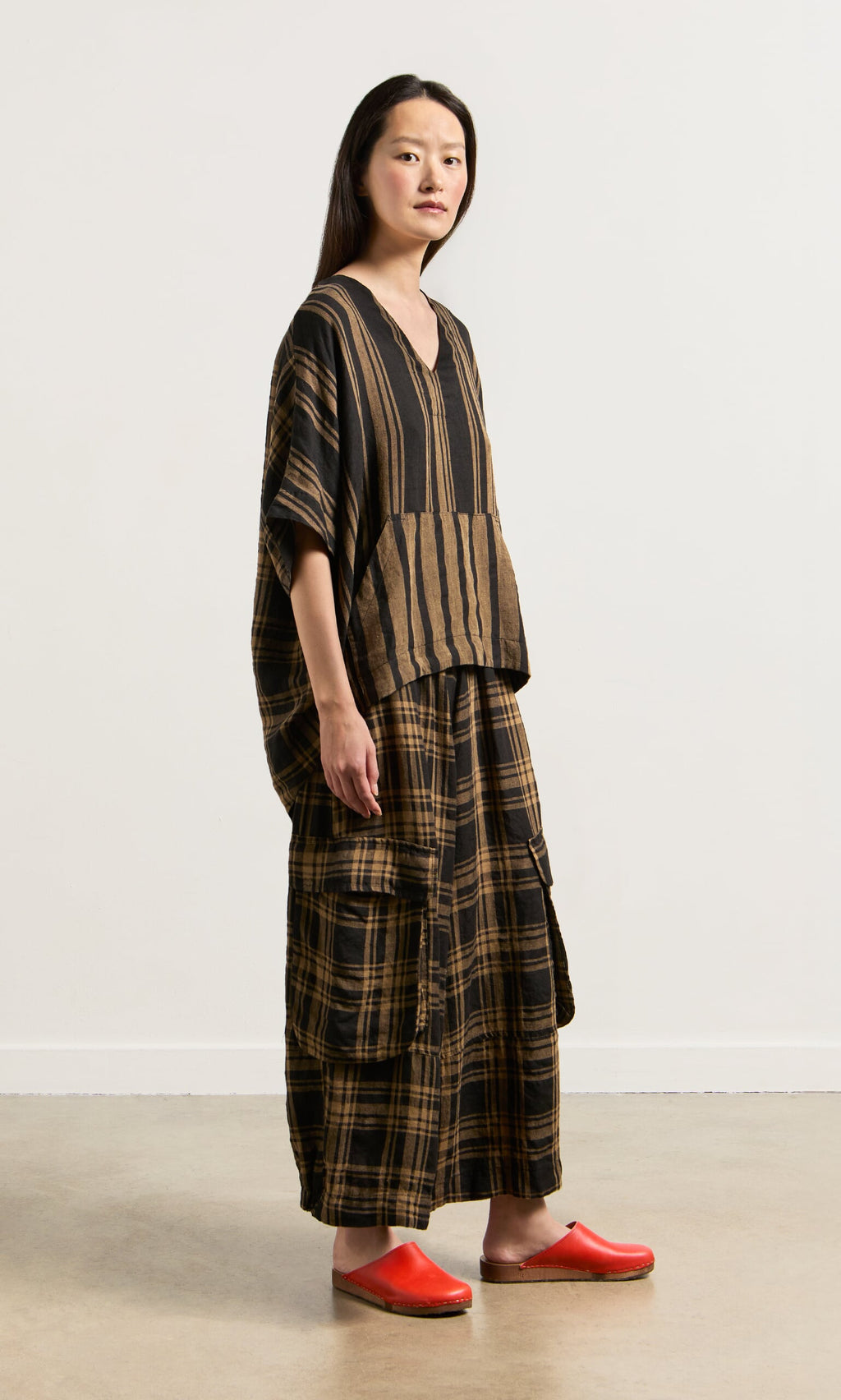 Plümo- Woman wearing the Westmorland Top, a loose V-neck with striped and checked panels, red clogs, indoors by a light wall.