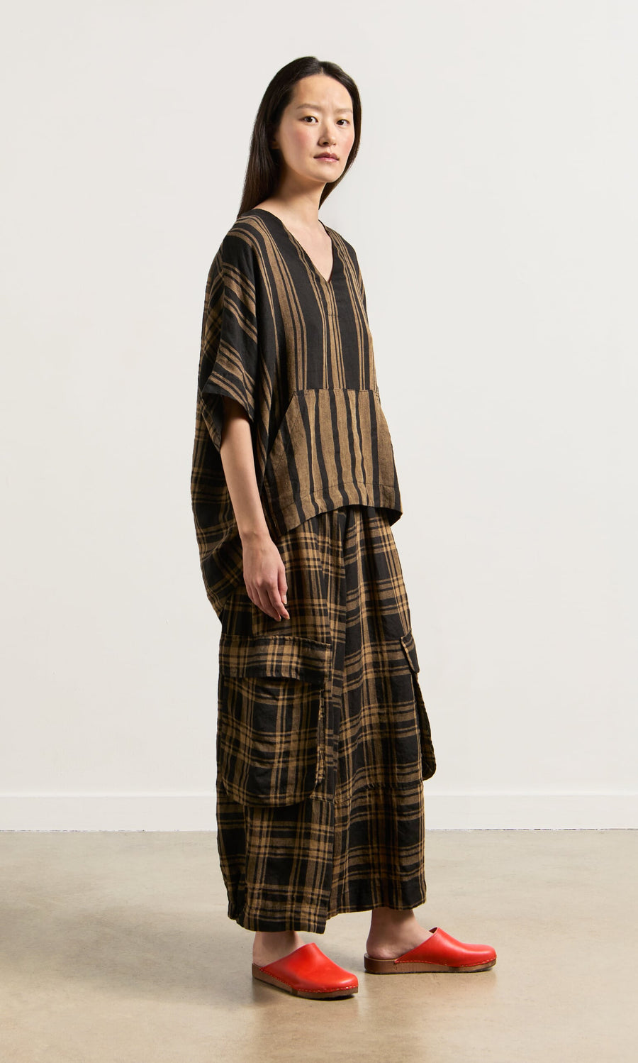 Plümo- Woman wearing the Westmorland Top, a loose V-neck with striped and checked panels, red clogs, indoors by a light wall.
