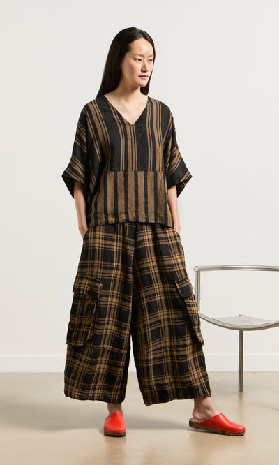 Plümo- Woman in a loose V-neck Westmorland Top with striped and checked panels stands by a metal chair.