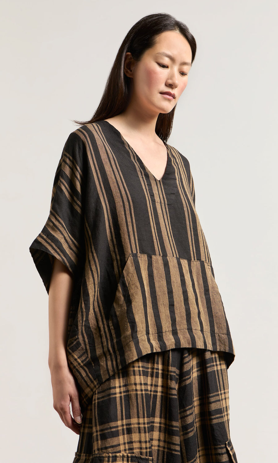 Plümo- Woman wearing the Westmorland Top—a loose woven linen V-neck with striped and checked panels, short sleeves, and a pocket.
