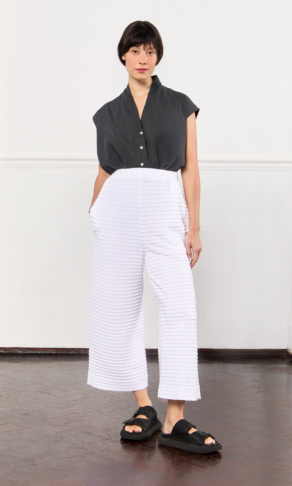 Plümo- Person in a black top, White Pintucked Pants, and black sandals, standing indoors against a white wall.