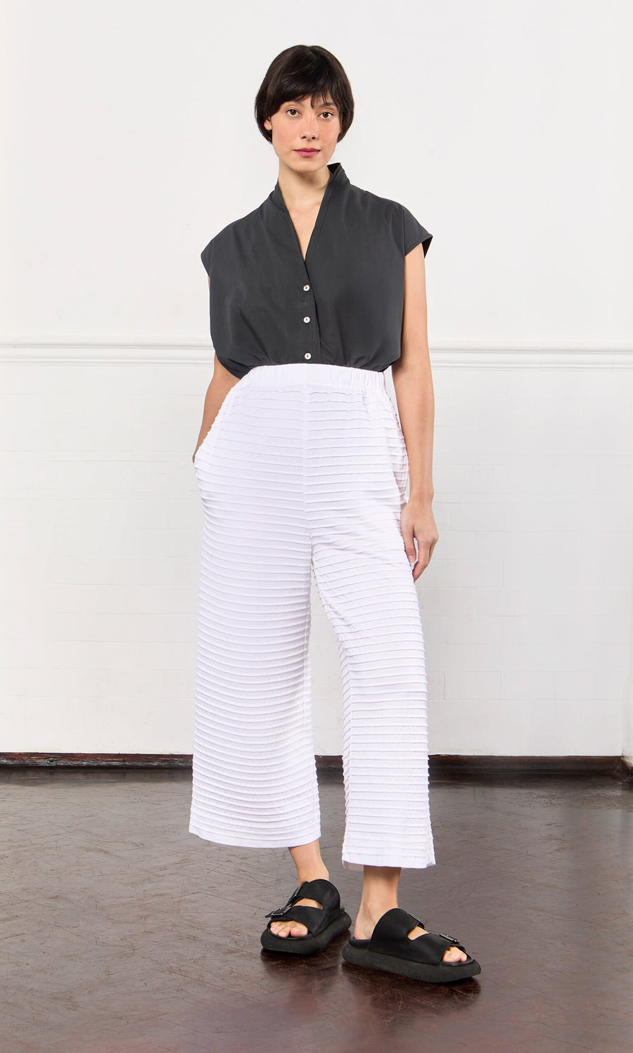 Plümo- Person in a black top, White Pintucked Pants, and black sandals, standing indoors against a white wall.