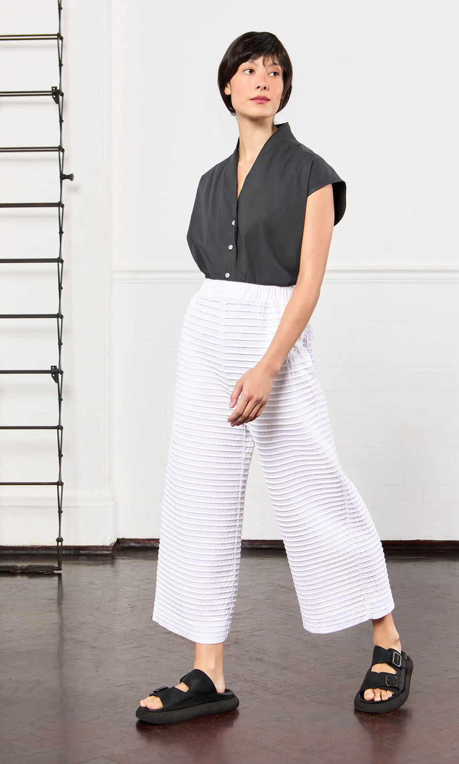 Plümo- A person in a black top and White Pintucked Pants stands indoors on a dark floor, looking to the side.