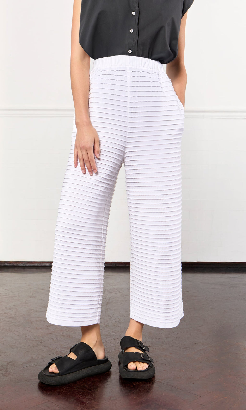 Plümo- Person wearing White Pintucked Pants, black sleeveless top, and black sandals, standing indoors.