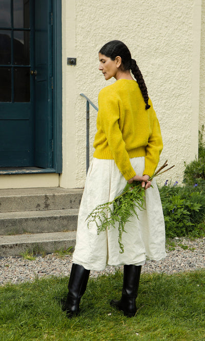 Plümo  Woman in Wicklow Crewneck - Golden Hop and white skirt holds greens behind her back by a blue door.