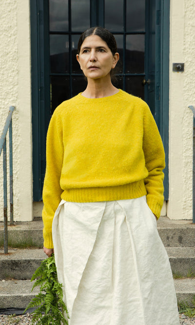 Plümo  Woman in a Golden Hop Wicklow Crewneck and white skirt stands outside, holding green leaves by a doorway.