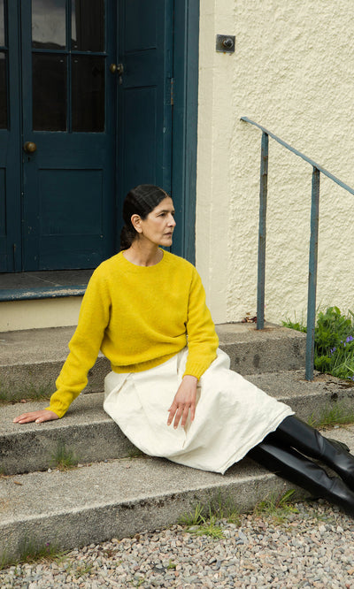 Plümo  Woman in a Wicklow Crewneck - Golden Hop sweater and white skirt sits on steps by a blue door, looking left.