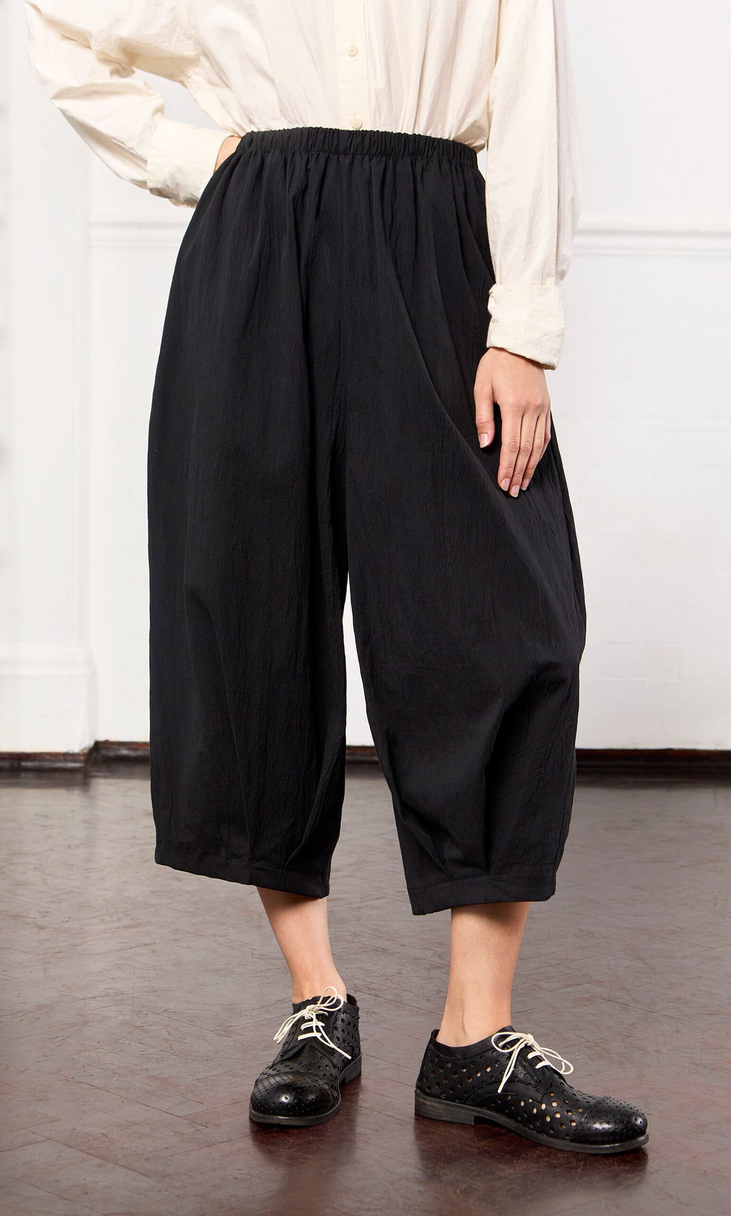 Plümo- Person wears Black Pinch Pants, a cream shirt, and black lace-up shoes while standing on a wooden floor.