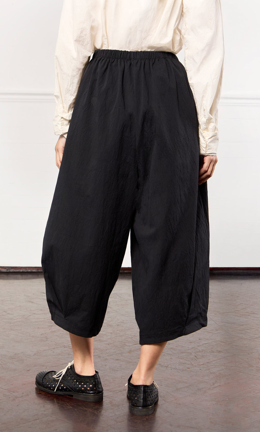 Plümo- Person wearing Black Pinch Pants with an elasticated waist, a cream top, and black shoes on a brown floor.