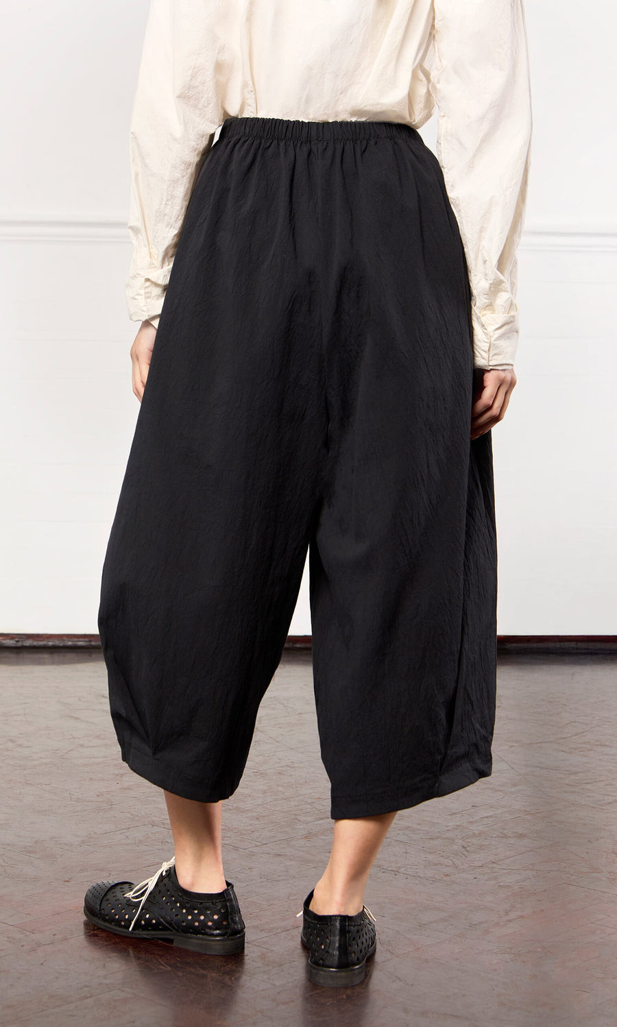Plümo- Person wearing Black Pinch Pants with an elasticated waist, a cream top, and black shoes on a brown floor.