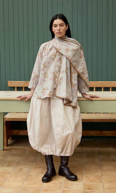 Plümo  Woman in floral top, scarf, and Edina Skirt - Nougat with elastic waist stands by a green wall and bench.