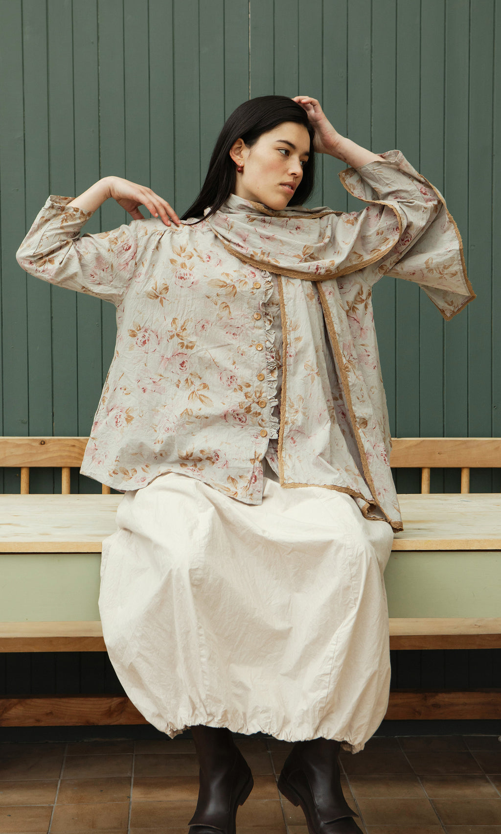 Plümo  A woman in the Dawn Scarf and a white skirt sits on a wooden bench against a green paneled wall.