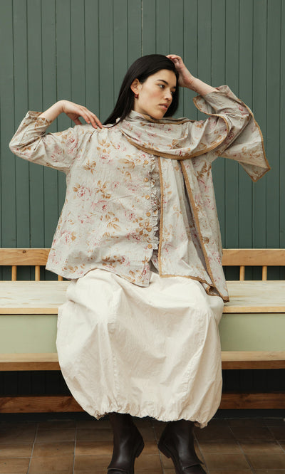 Plümo  A woman in the Dawn Scarf and a white skirt sits on a wooden bench against a green paneled wall.