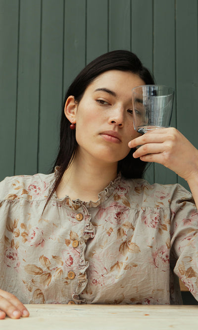 Plümo  Woman wears the Wild Flower Blouse, its ruffled trims and vintage florals framing her thoughtful gaze behind an empty glass.