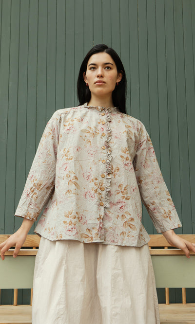 Plümo  Woman in the Wild Flower Blouse with ruffled trims and white skirt stands before a green paneled wall, looking ahead.