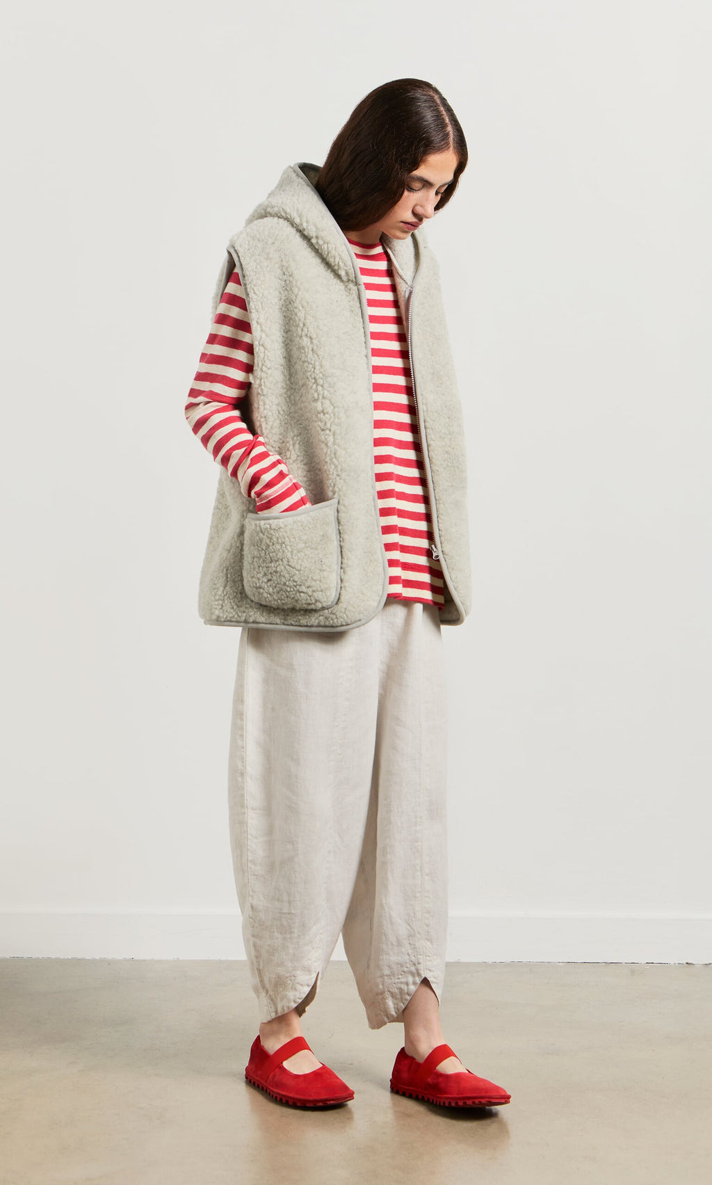 Plümo- Person in a red and white striped top, Wool Gilet - Light Grey, loose white pants, and red shoes, standing and looking down.