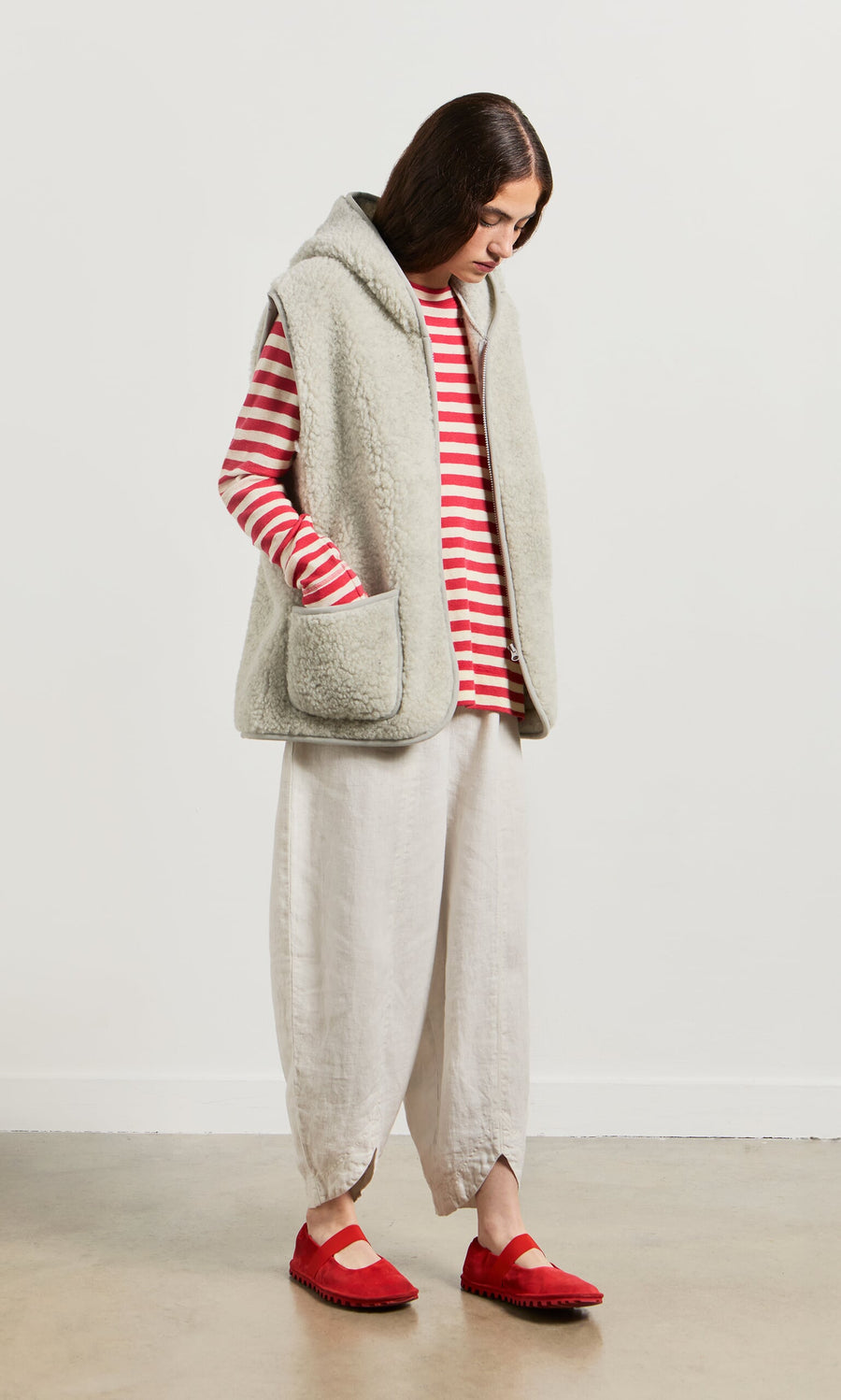 Plümo- Person in a red and white striped top, Wool Gilet - Light Grey, loose white pants, and red shoes, standing and looking down.