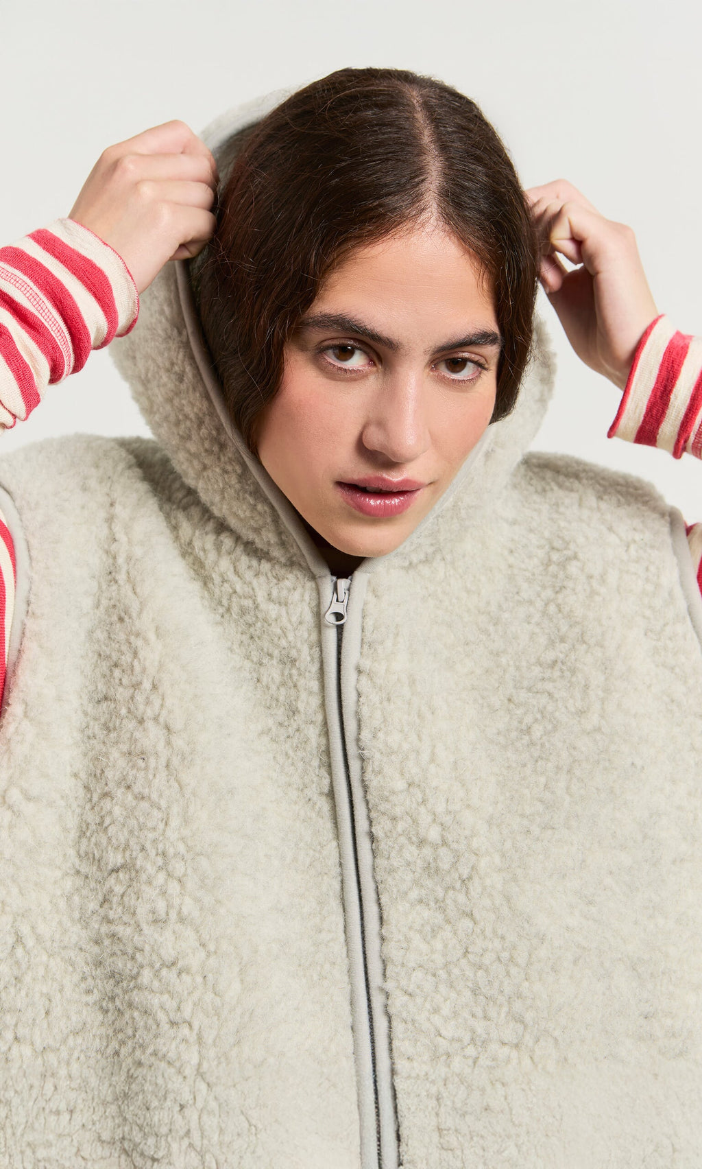 Plümo- Woman in a light grey Wool Gilet, zipping up over a pink and white striped long-sleeve shirt.