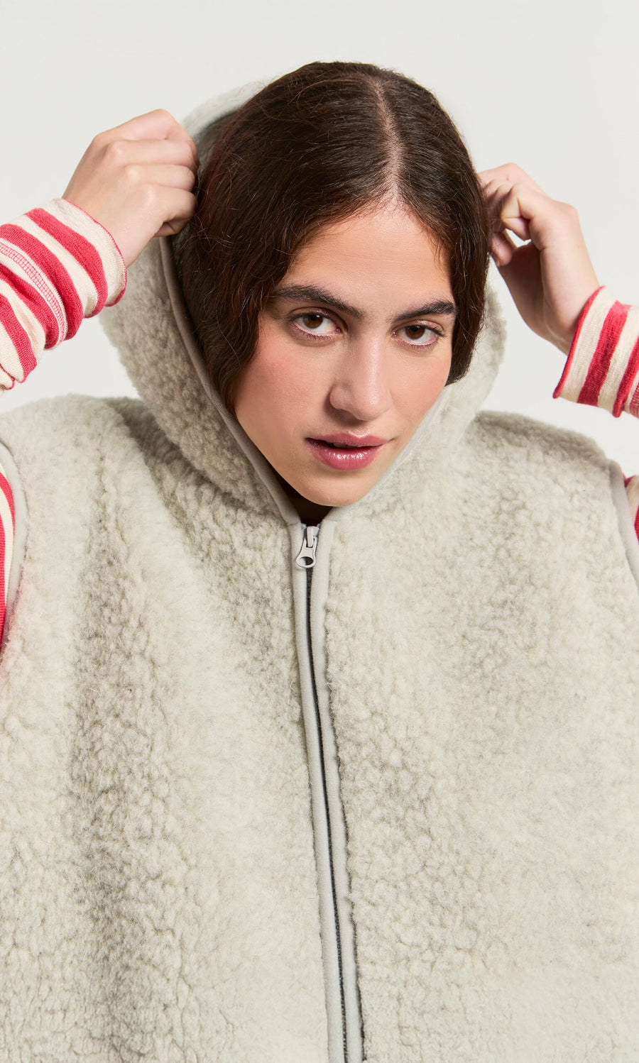 Plümo- Woman in a light grey Wool Gilet, zipping up over a pink and white striped long-sleeve shirt.