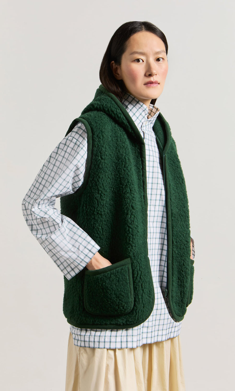 Plümo- A person wears the Wool Gilet - Moss Green over a checked shirt and cream skirt, standing against a plain background.