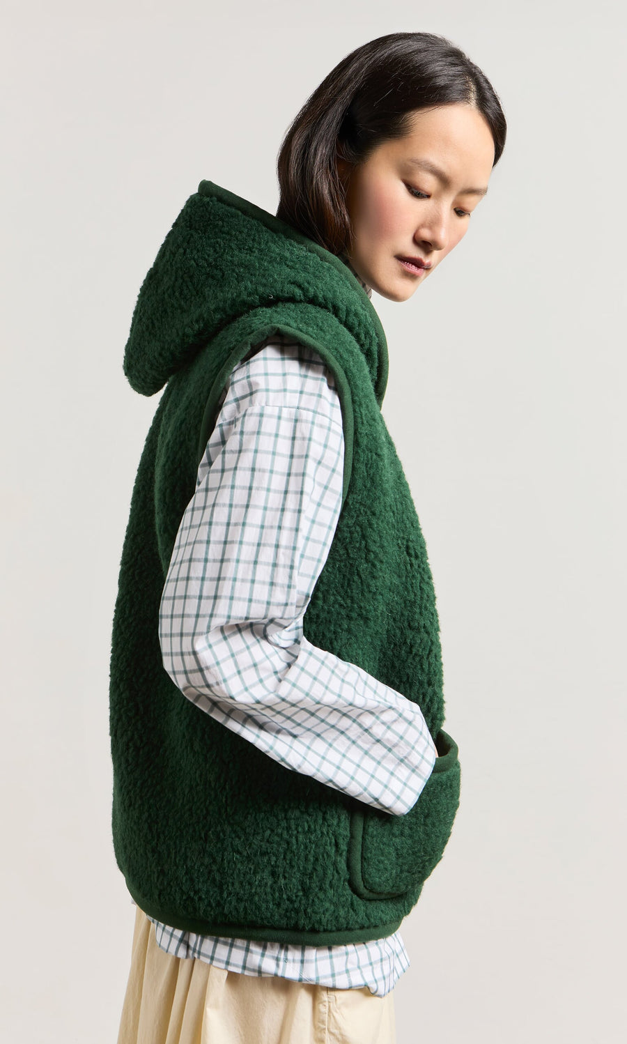 Plümo- Woman wearing a Moss Green Wool Gilet over a white checkered shirt, standing sideways with hands in pockets.