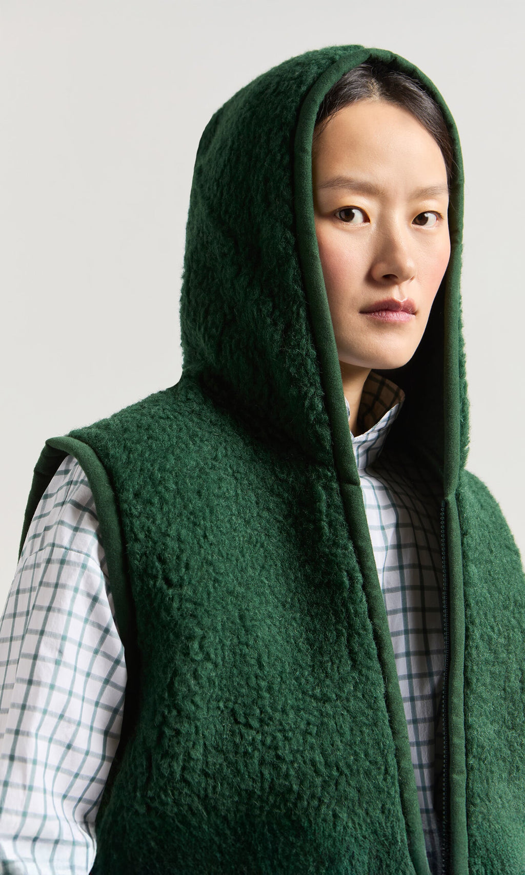 Plümo- Person wearing a Wool Gilet - Moss Green over a plaid shirt, looking at the camera with a neutral expression.
